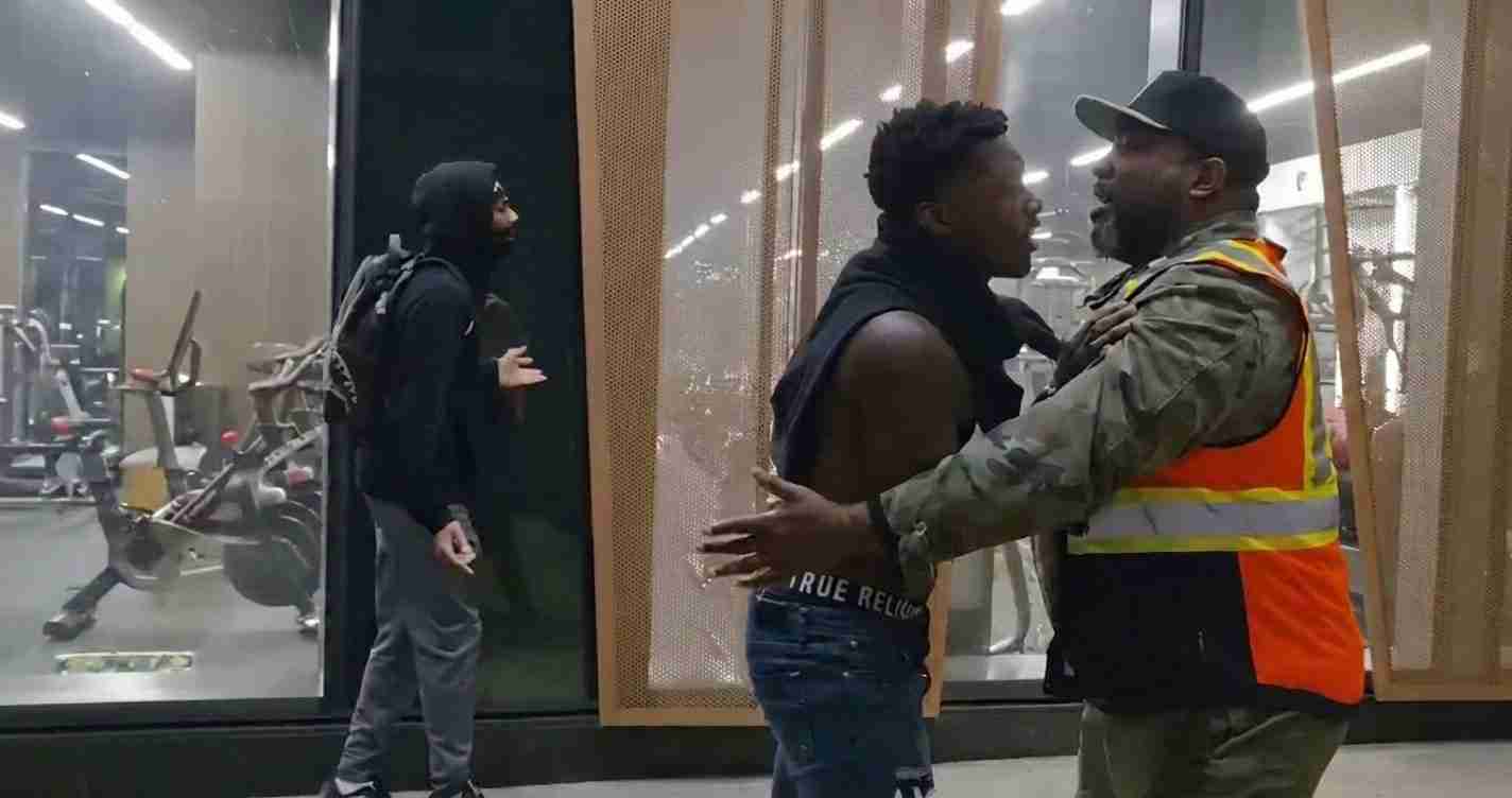 Security guard in an orange vest restrains a shirtless man near a gym glass wall while a masked bystander in a hoodie watches in the background.