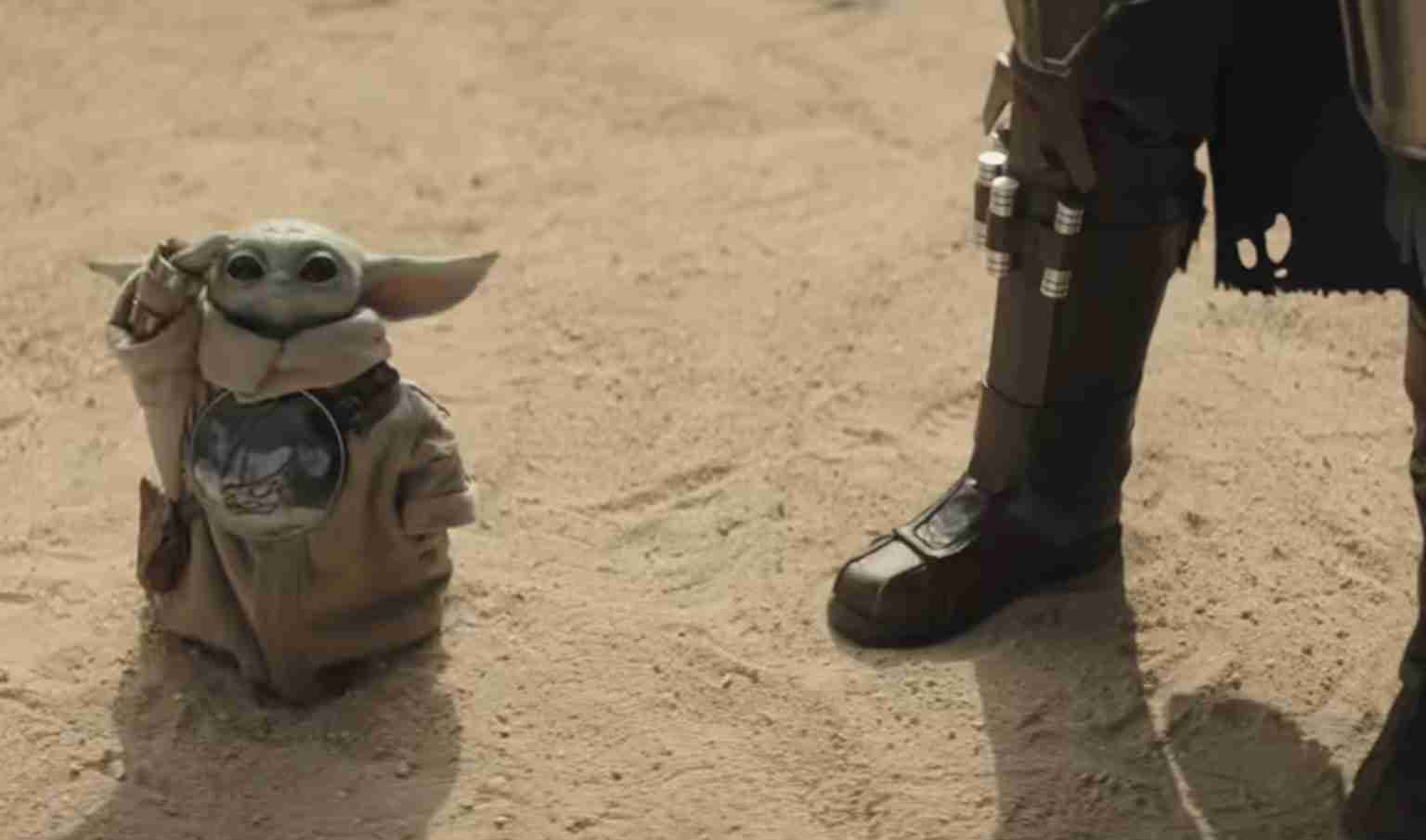 Grogu (Baby Yoda) sits in a tan sack on a dusty desert ground, peeking out while a armored boot stands nearby.