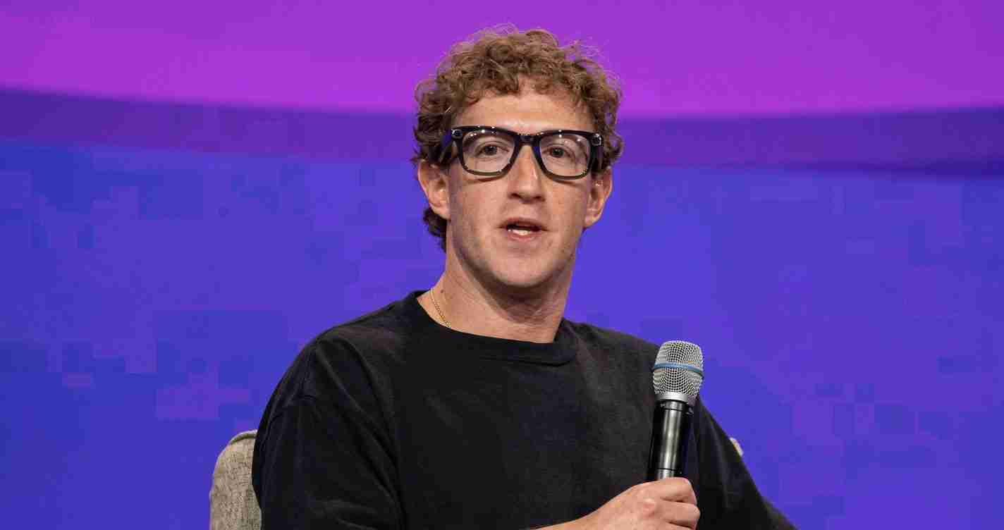 Man wearing a black shirt and dark glasses speaks into a handheld microphone on a purple stage backdrop.
