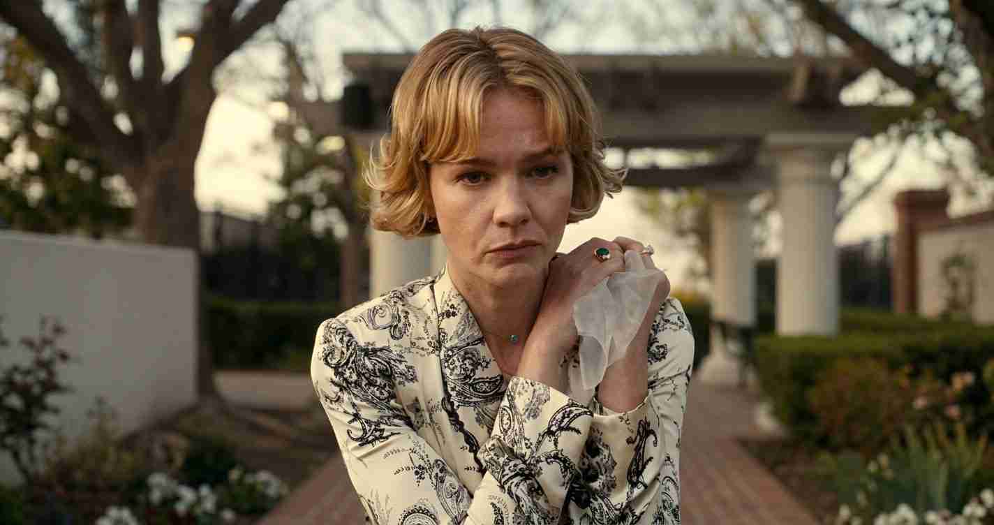 Woman with short blond hair in a patterned blouse stands outdoors, clutching a tissue and looking pensive in a garden setting.