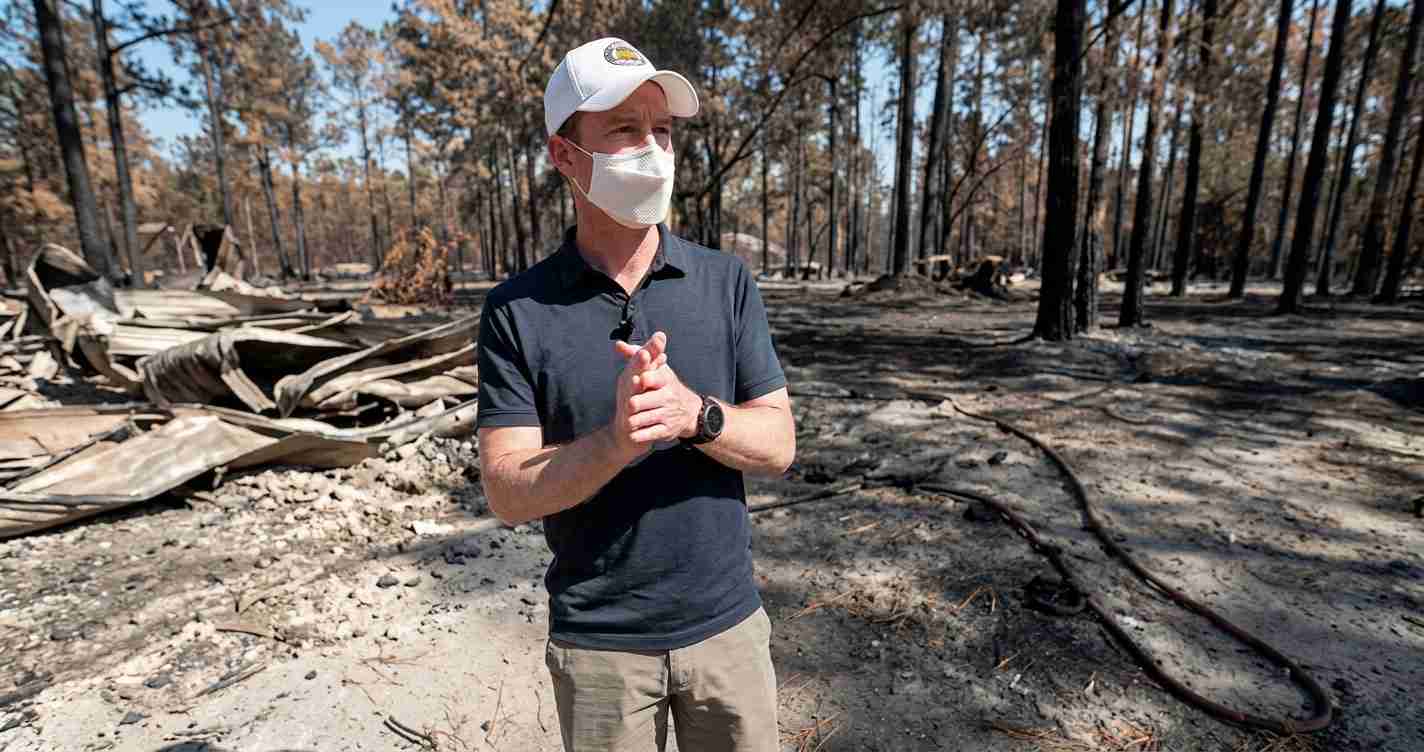 Man wearing a mask and cap stands amid a burned forest with charred trees and debris scattered on the ground.