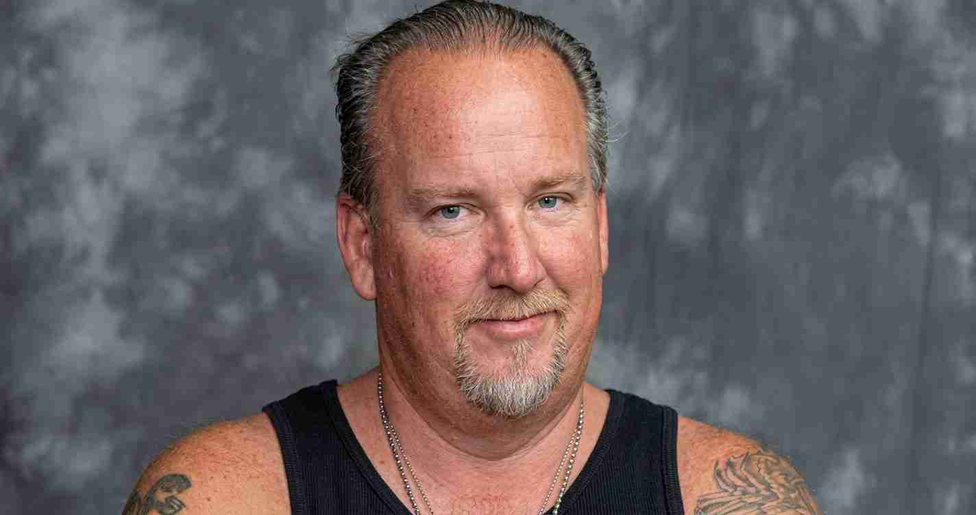 Man with short hair and a goatee wearing a black sleeveless shirt, tattoos on both arms, posed against a gray studio backdrop.