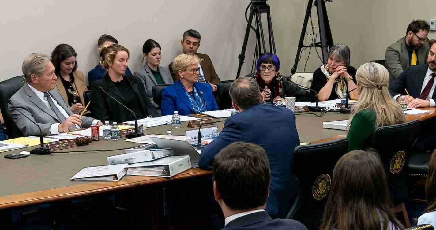 Diverse group of officials seated around a long conference table during a formal meeting, with microphones, binders, and water bottles nearby.