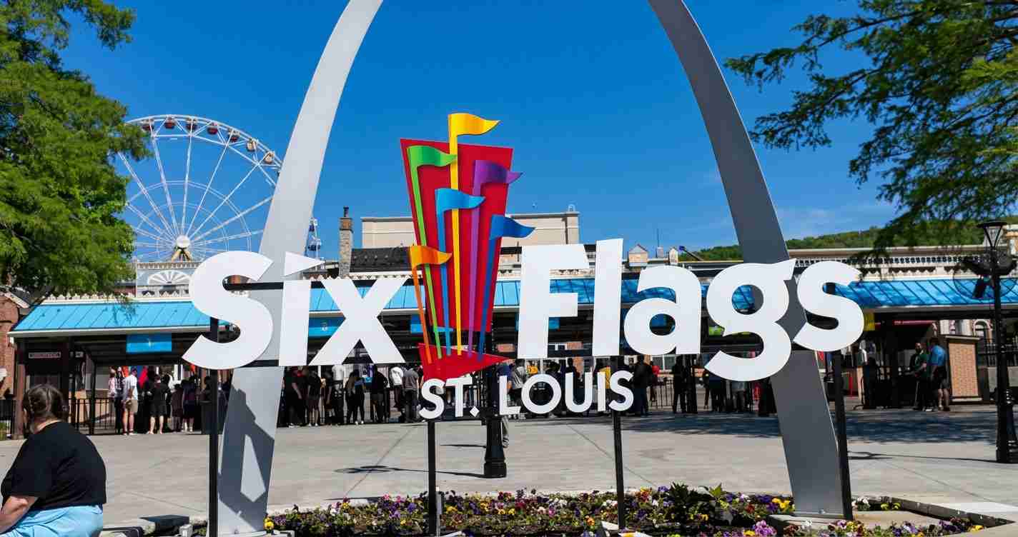 Six Flags St. Louis entrance sign with a colorful flag sculpture and a Ferris wheel in the background, with visitors nearby.
