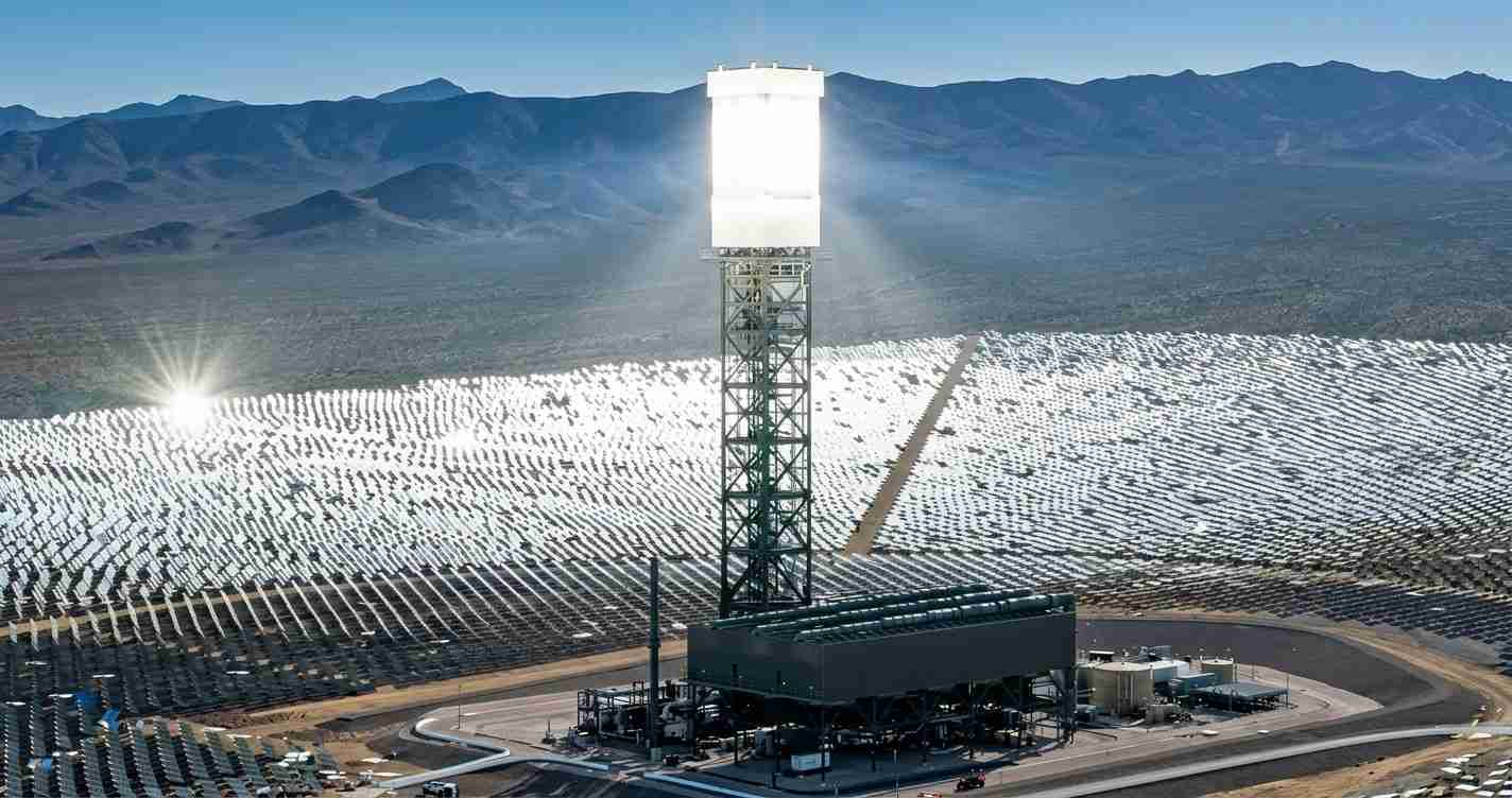 Solar power plant with a tall central receiver tower and rows of mirrors spread across the desert. Mountains rise in the background.
