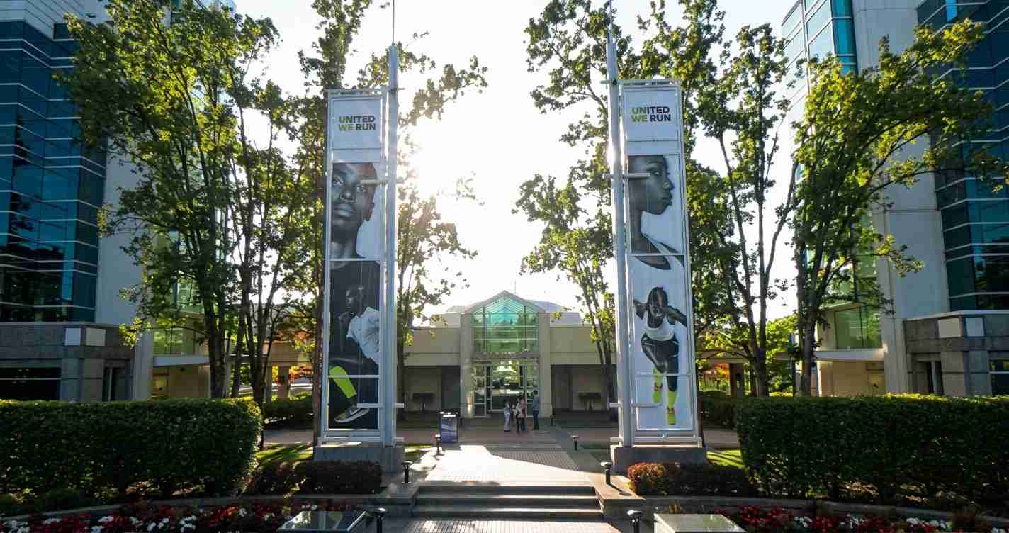 Outdoor plaza in front of a modern office complex; two vertical banners reading 'United We Run' flank a glass entrance as sunlight filters through surrounding trees.