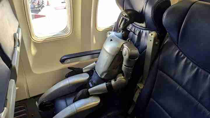 Small silver humanoid robot seated in an airplane window seat, arms folded over its lap, looking out the window.