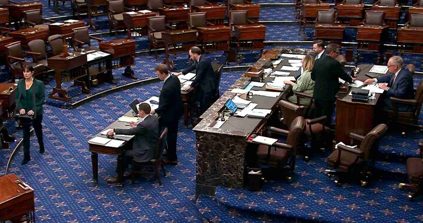 Legislative chamber with desks, a marble podium, and several people in suits working with papers and laptops around the room.