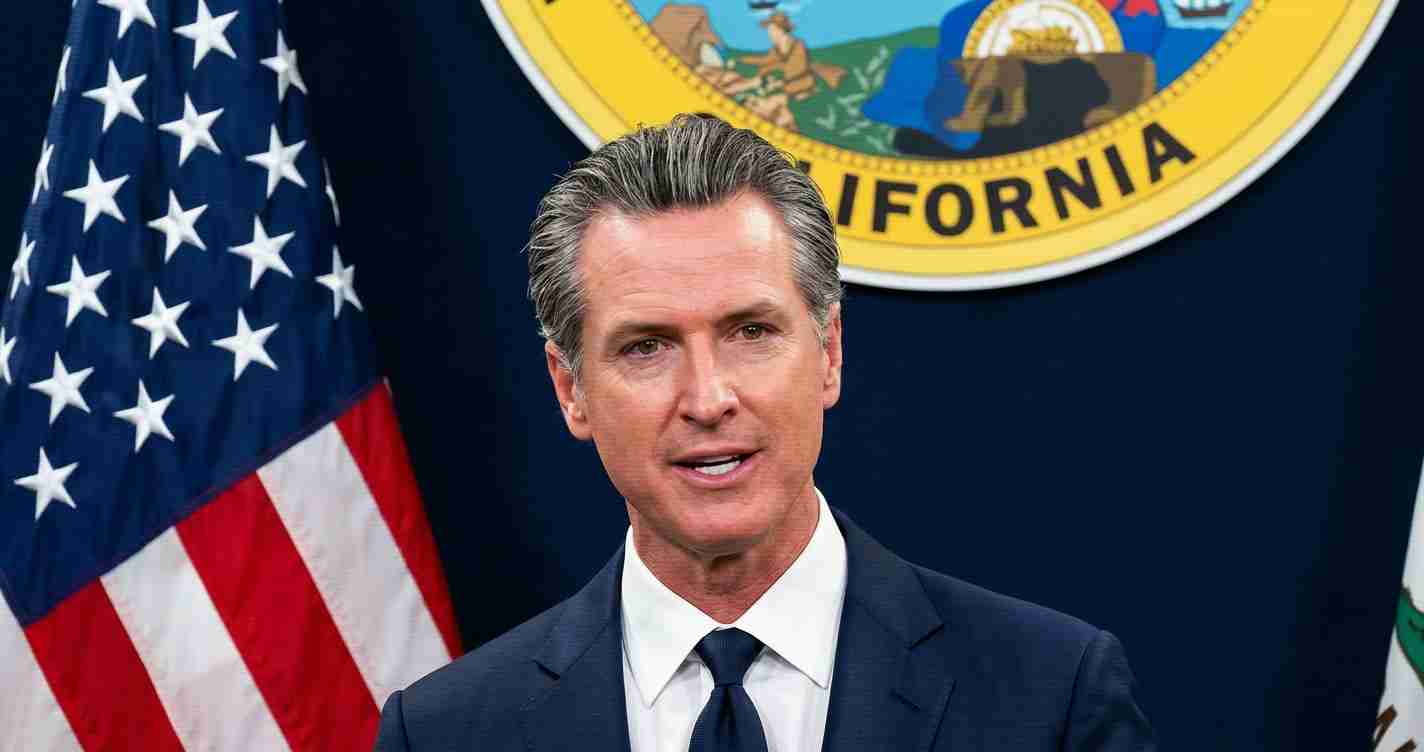 Man in a dark suit and tie speaking at a press briefing, with the American flag and a state seal visible in the background.