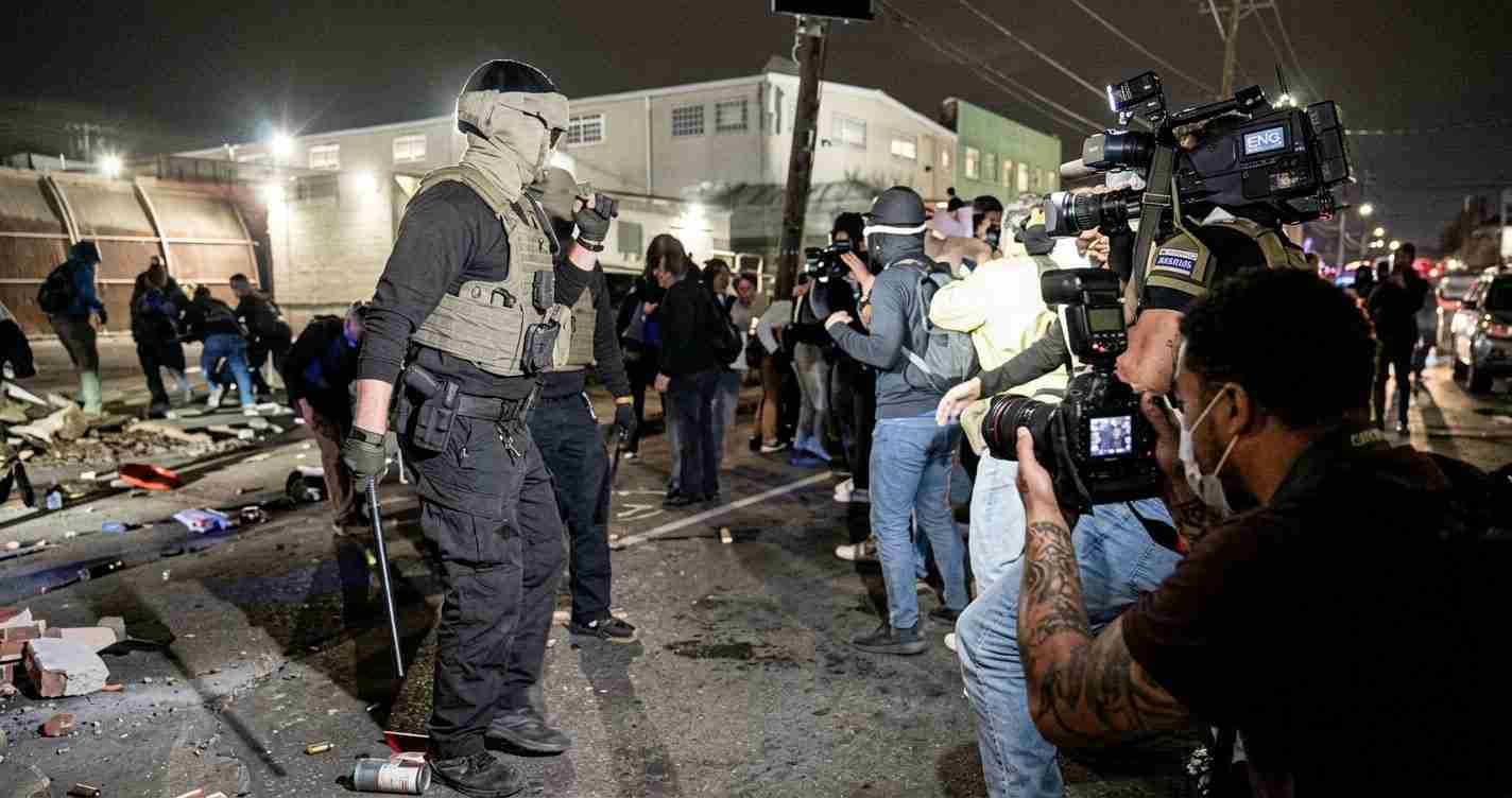 Nighttime street scene with a heavily armed officer in tactical gear facing a line of photographers filming the scene.
