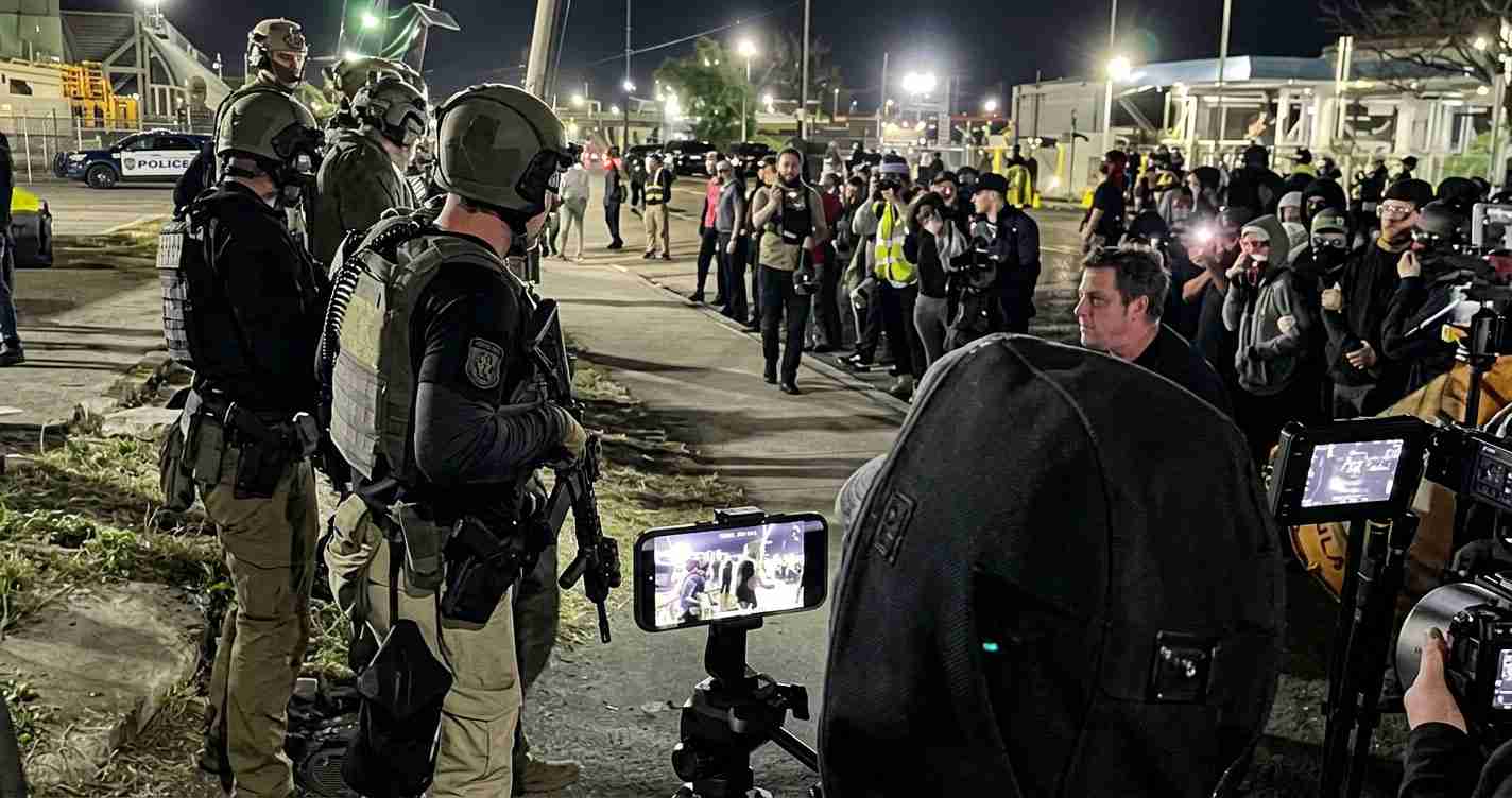 Night scene of heavily armed soldiers in tactical gear facing a crowd of protesters and journalists, with cameras and phones recording the moment.
