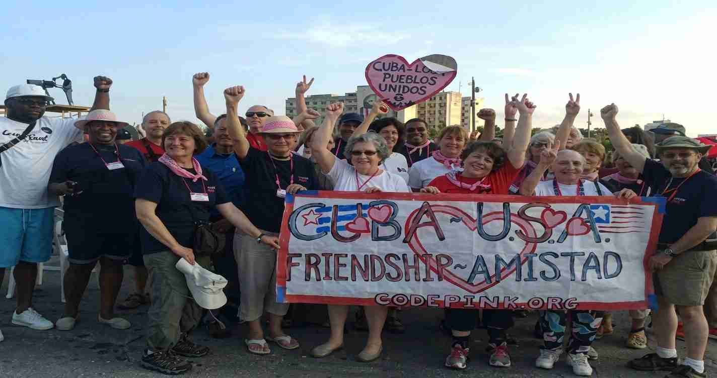Group of diverse people at a Cuba–USA friendship event, holding a banner that reads Cuba–USA Friendship/Amistad with heart motifs on the sides.