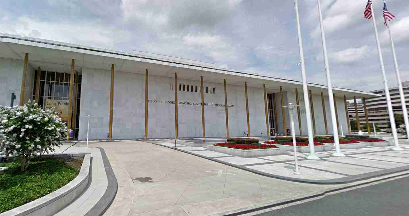 Exterior view of a modern memorial building with a row of gold columns, flagpoles, and landscaped plaza in front.