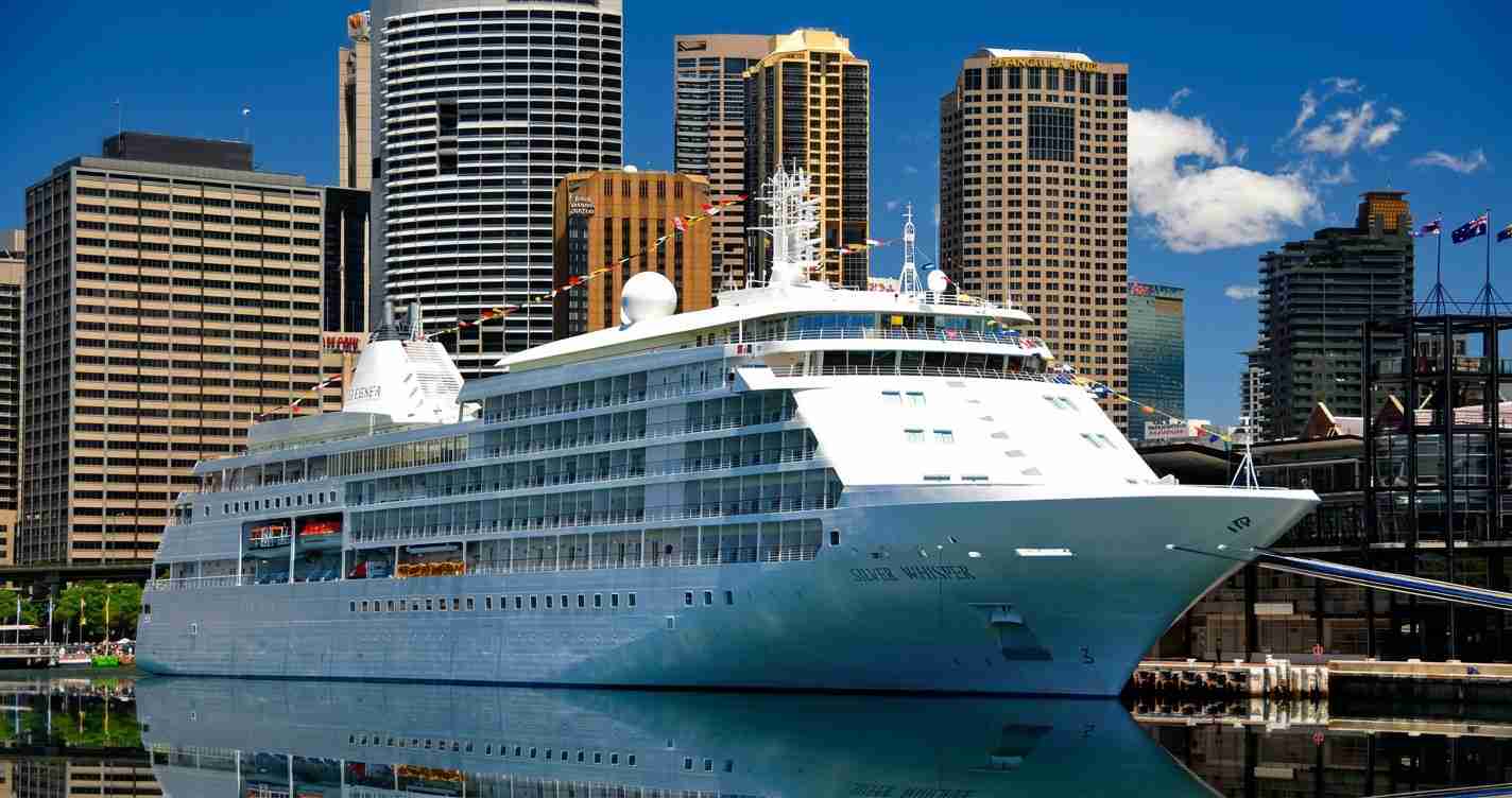 White cruise ship docked at a modern city harbor with tall buildings in the background.