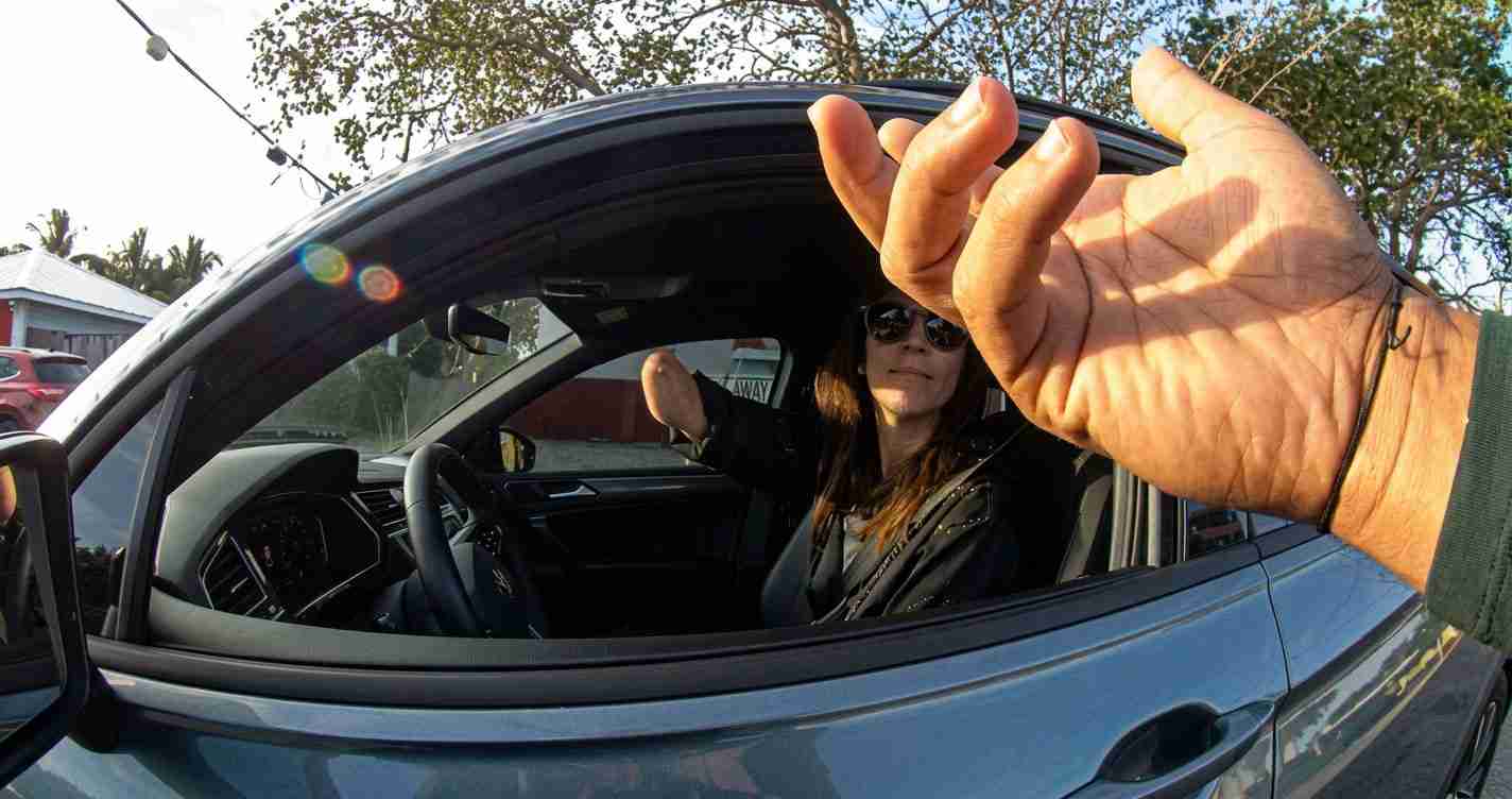 Woman driving a blue car leans out with her arm toward the camera as the driver’s window is open; sun glare visible.