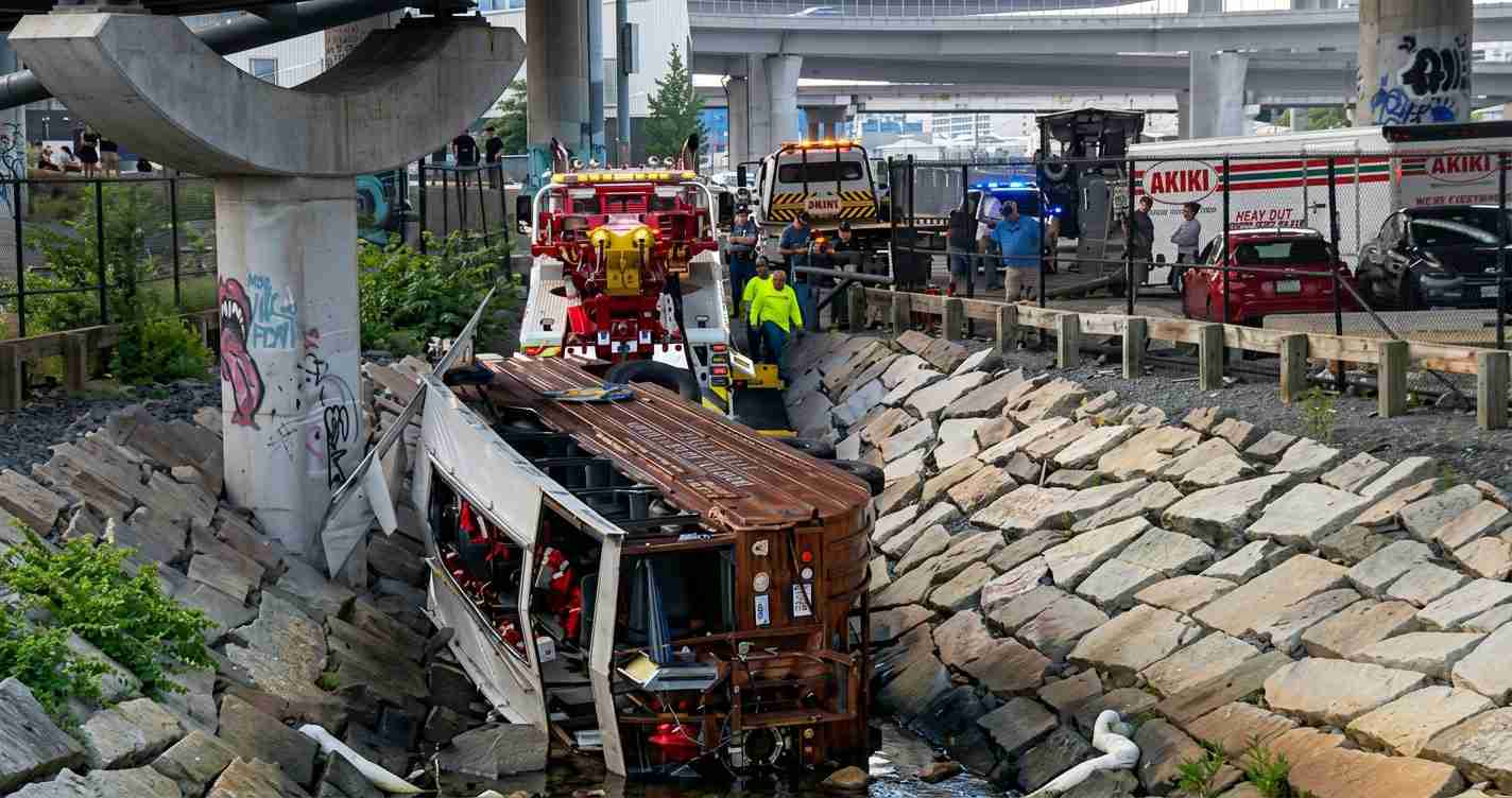 Emergency responders inspect a derailed bus wedged in a rocky drainage ditch beneath an overpass.