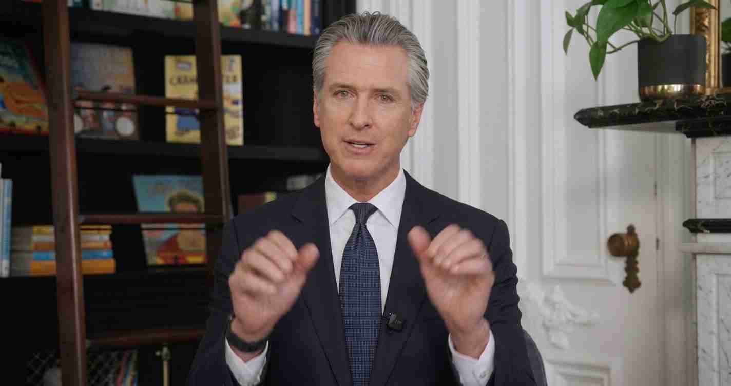 Man in a dark suit and tie speaks to camera, hands raised mid-gesture, with bookshelves behind on the left.
