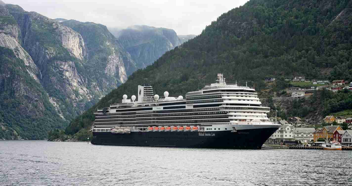 Large black-and-white cruise ship anchored near a fjord village with cliffs and green hills in the background