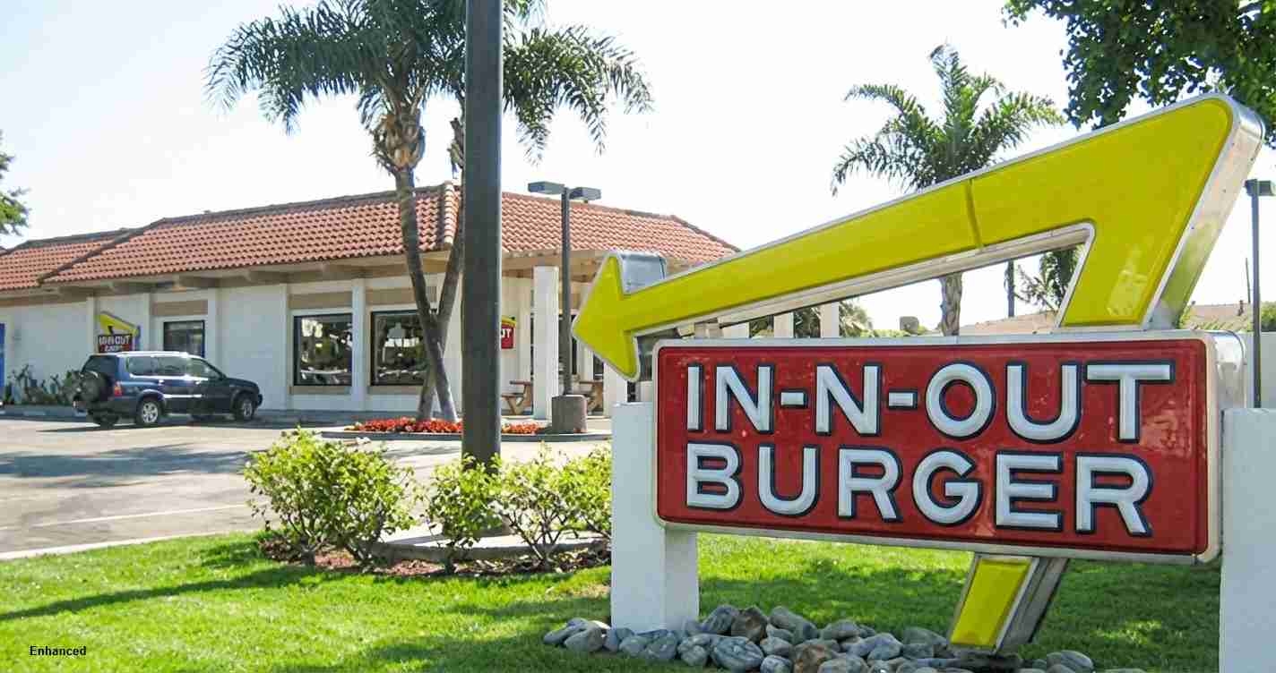 Large retro In-N-Out Burger sign with a bright yellow arrow in front of a fast-food building and palm trees.