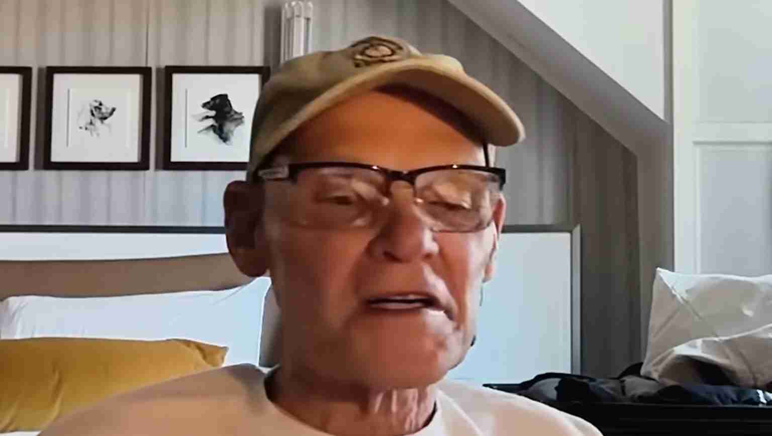 Older man wearing a beige cap and glasses speaks during a video call in a bedroom setting.