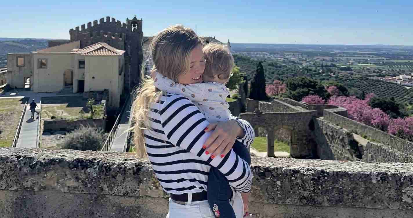 Woman in a striped top holds a toddler on a stone rampart with a historic fortress and valley views behind them.