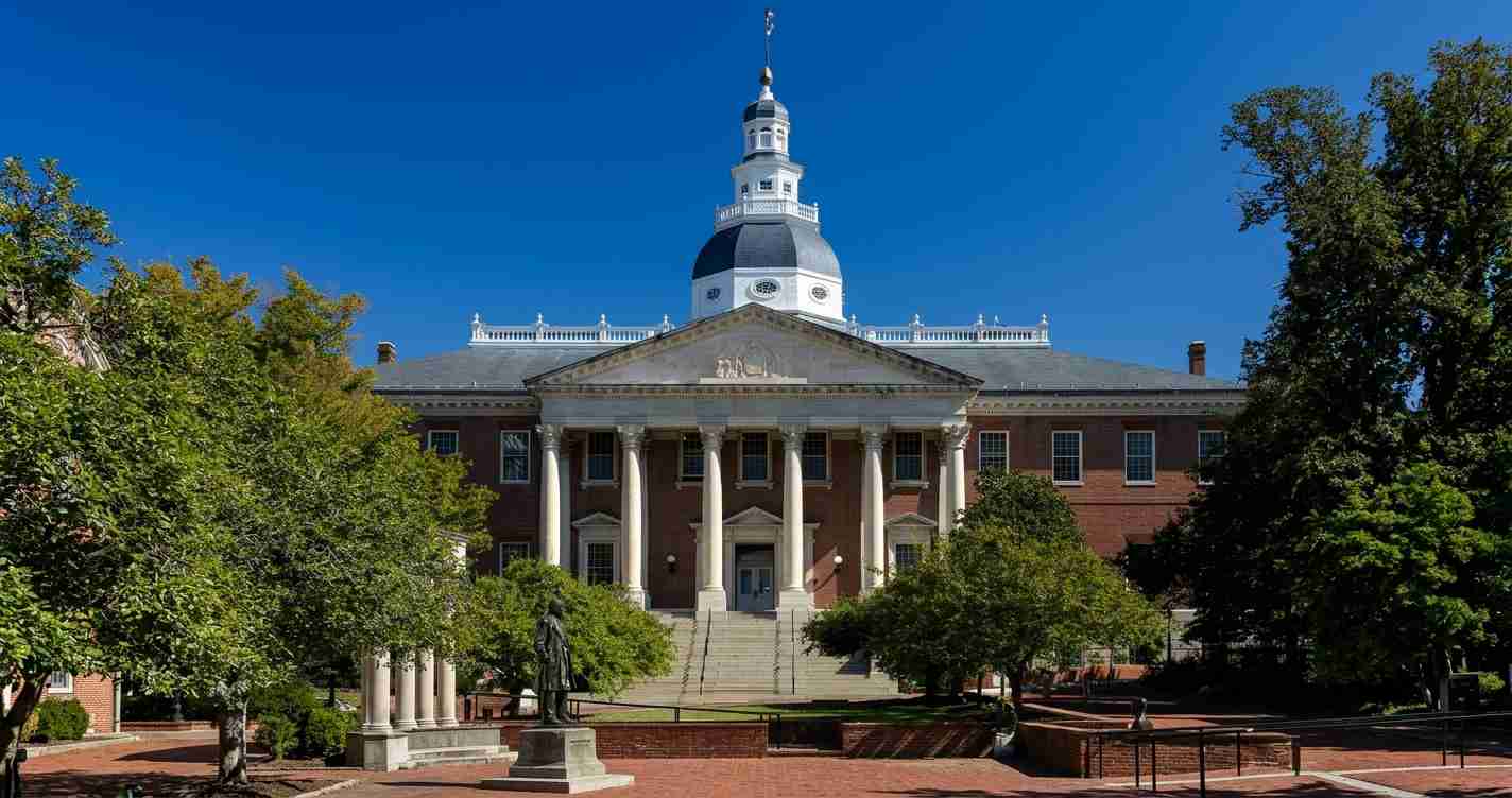 Front view of a brick university building with a white columned portico and a domed cupola atop the roof, set in a sunny plaza with trees and a statue nearby.