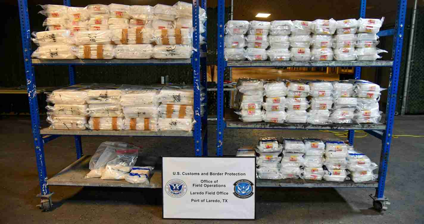 Stacks of white bags on blue metal shelves in a warehouse; a sign in front reads U.S. Customs and Border Protection, Port of Laredo, TX.