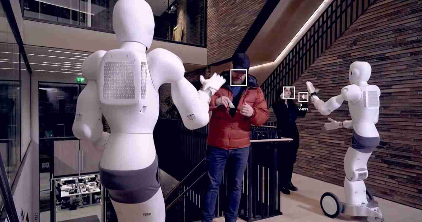 Two white humanoid robots extend their arms toward a man in a red jacket in a modern lobby or atrium.
