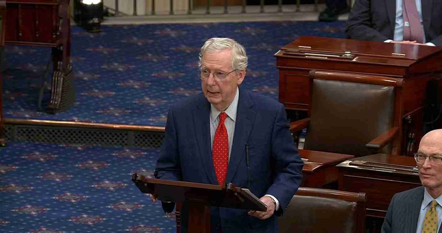 Older man in a navy suit and red polka-dot tie standing at a wooden podium in a legislative chamber, delivering a speech.