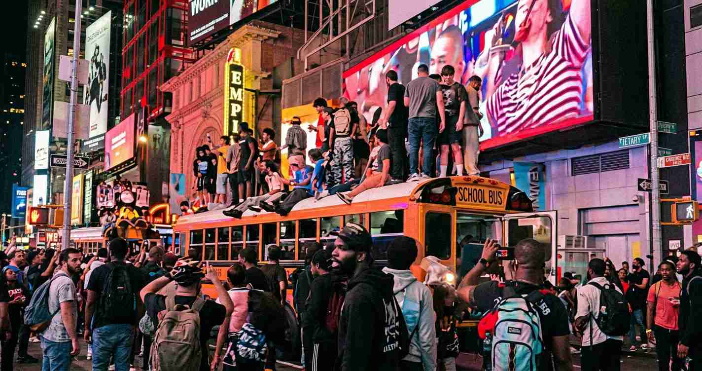 Crowd gathers around an orange school bus on a busy neon-lit city street, with people sitting and standing on the bus roof amid large billboards and screens.