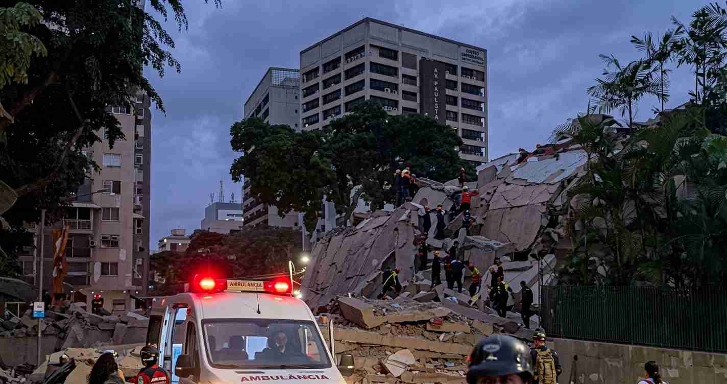 Emergency responders on a rubble-strewn collapsed building with an ambulance and flashing red lights in the foreground at dusk