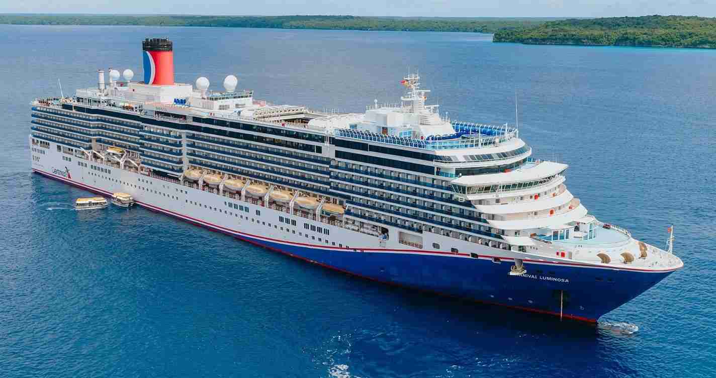 Large Carnival Cruise Line ship Carnival Luminosa sailing in calm blue water with multiple decks and lounges visible from above.