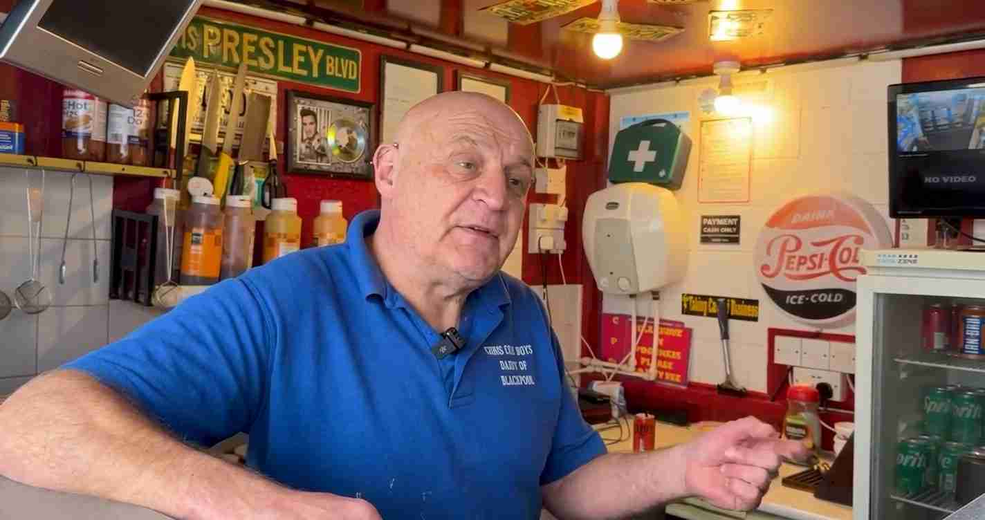 A bald man in a blue polo behind a counter in a small shop, gesturing as he speaks to a customer with signs and shelves in the background.