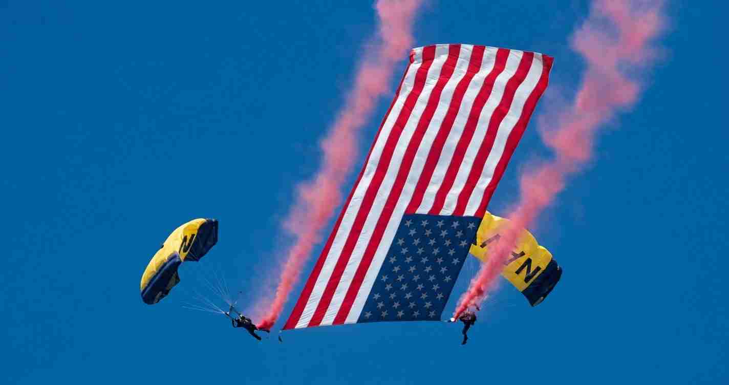 Two skydivers hold up a large American flag banner in midair against a clear blue sky, with pink smoke trails behind them.