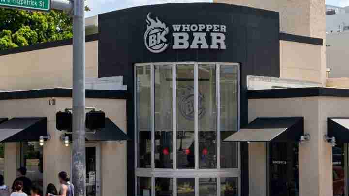 Whopper Bar storefront with a large BK logo, tall glass windows, and beige walls with black trim; pedestrians on the sidewalk nearby on a sunny day.