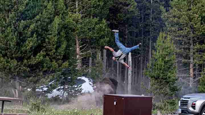 Person flipping through the air outdoors near a dusty demolition, with trees and parked cars in the background.