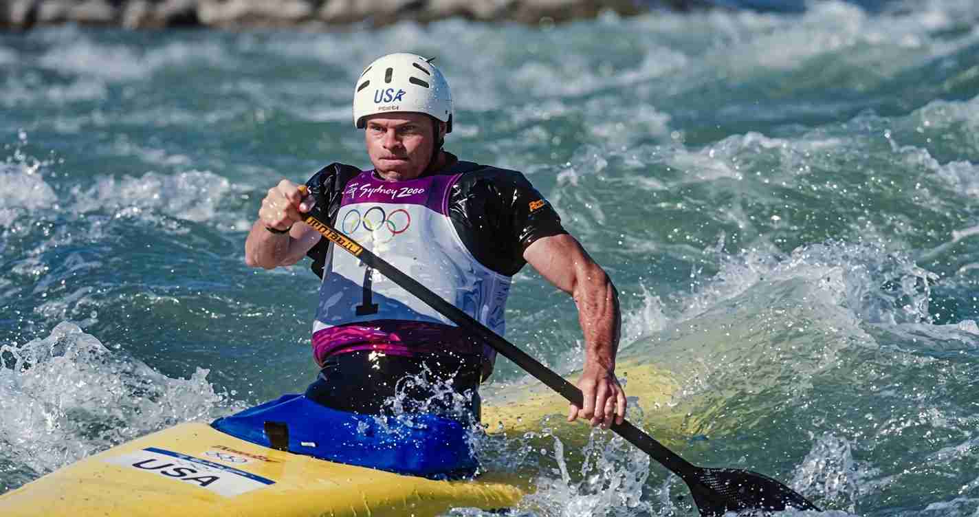 Male kayaker in a white helmet paddling through choppy water in a yellow USA kayak, wearing Olympic gear.