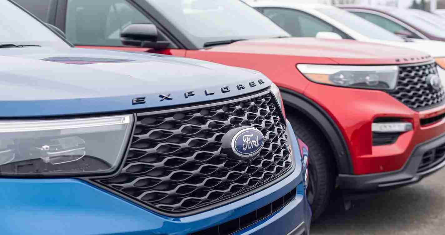Close-up of Ford Explorer grilles on blue and red SUVs in a dealership lot, emphasizing the brand badge and honeycomb grills, row of cars behind them