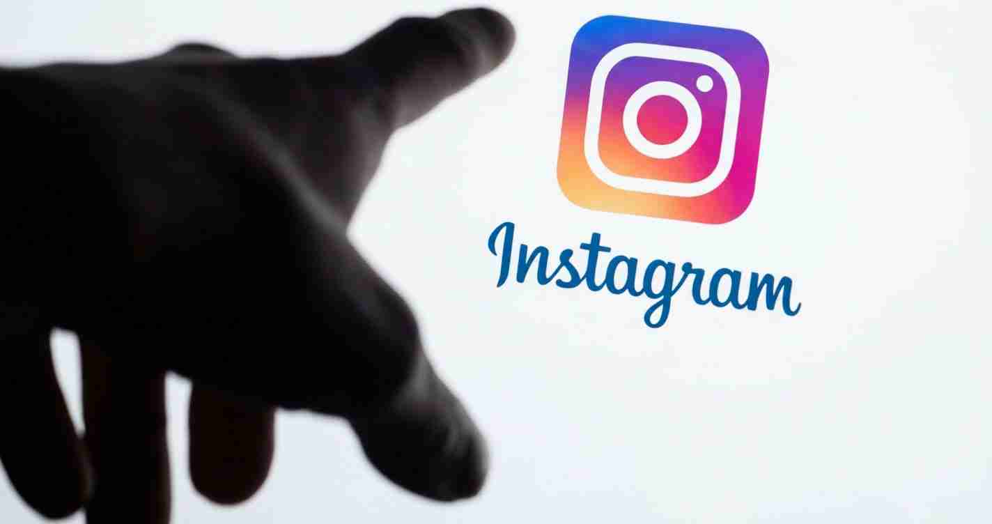 Hand reaching toward the Instagram logo on a light background, suggesting social media interaction