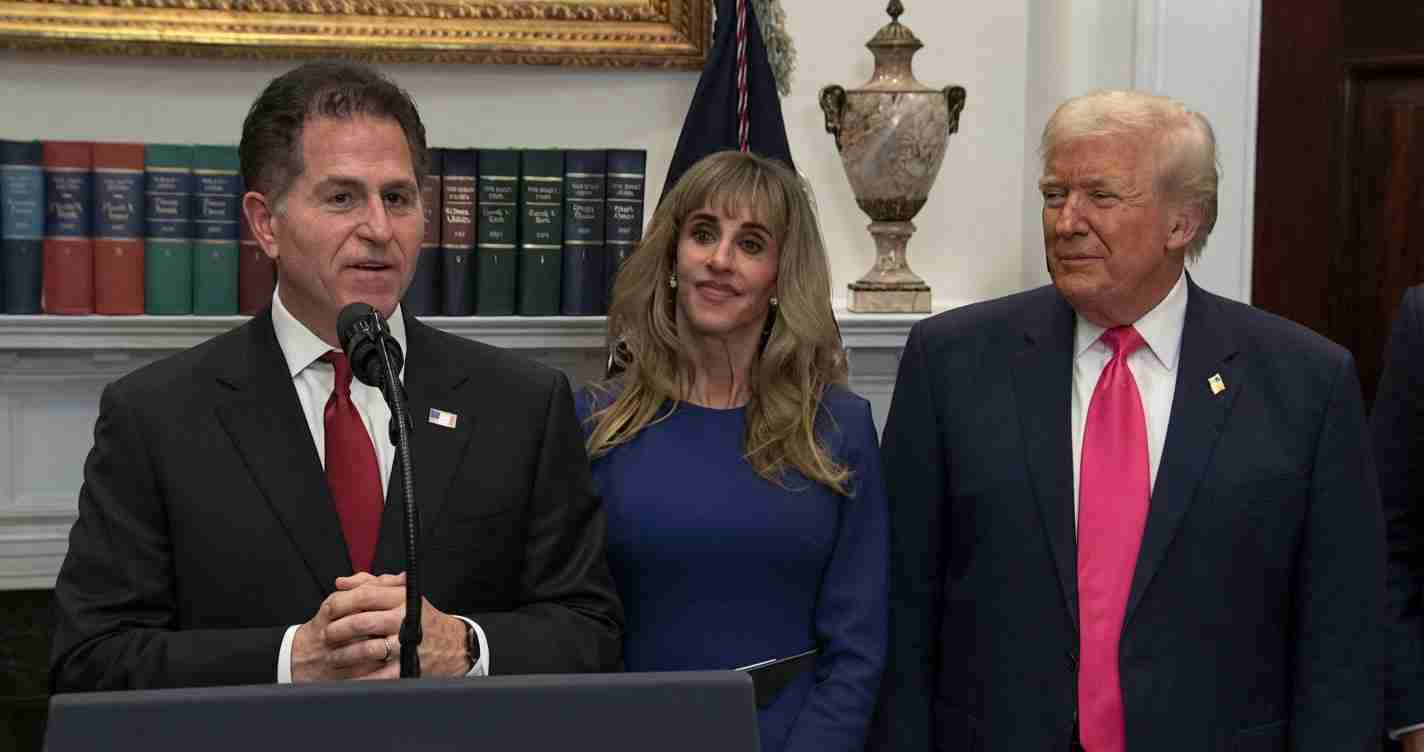 Man in a dark suit and red tie speaks at a podium with a microphone, flanked by a woman in a blue dress and a man in a dark suit with a pink tie in a formal room with books and a decorative urn behind them.