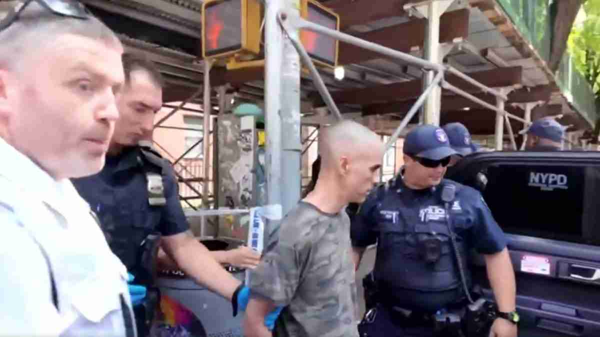 Man in camouflage shirt stands with hands behind back as officers detain him beside a black NYPD SUV under a construction scaffold area.