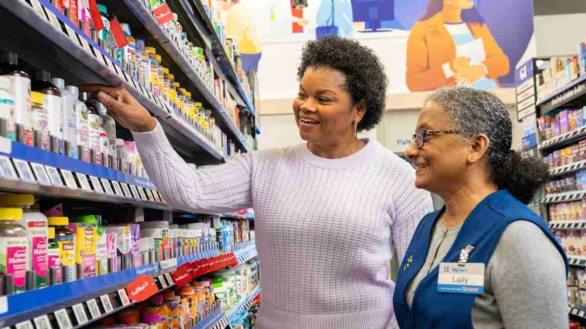 Smiling shopper reaches for a bottle on a pharmacy aisle shelf as a store associate in a blue vest looks on approvingly.