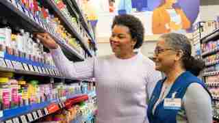 Smiling shopper reaches for a bottle on a pharmacy aisle shelf as a store associate in a blue vest looks on approvingly.