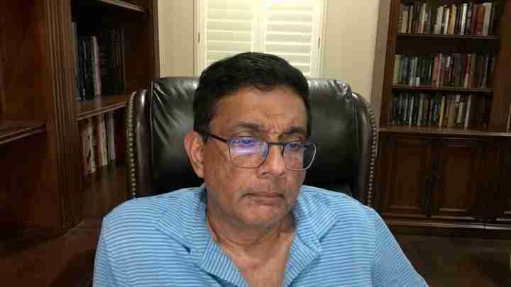 Head-and-shoulders of a man wearing glasses, seated in a leather chair in a home library, wearing a light blue striped polo shirt.