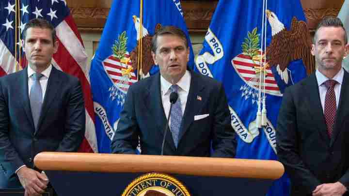 Three men in suits stand at a podium with DOJ flags behind them, during a press conference. The center speaker addresses the room while two aides stand beside him.